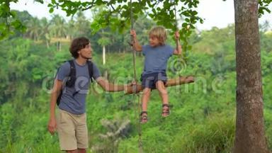 一个年轻人在热带公园的秋千上摆动他的小儿子的慢镜头。 儿童概念